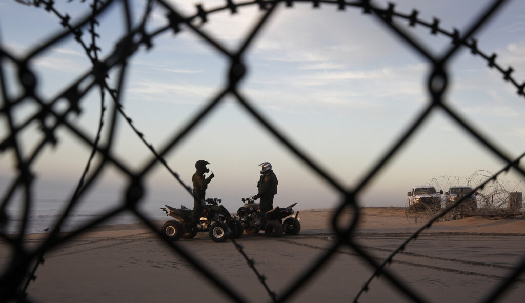 Two U.S. Border Patrol agents talk along the beach in San Diego, Wednesday, Jan. 9, 2019, seen through razor wire lining the border wall from Tijuana, Mexico.