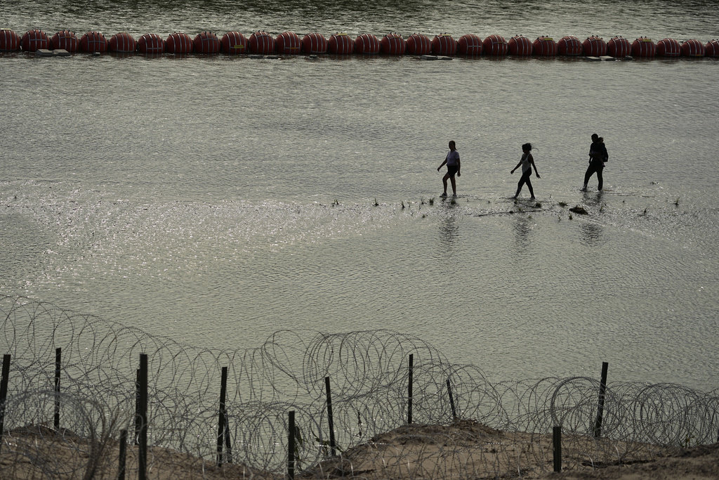 Immigrants walk past large buoys being used as a floating border barrier on the Rio Grande.