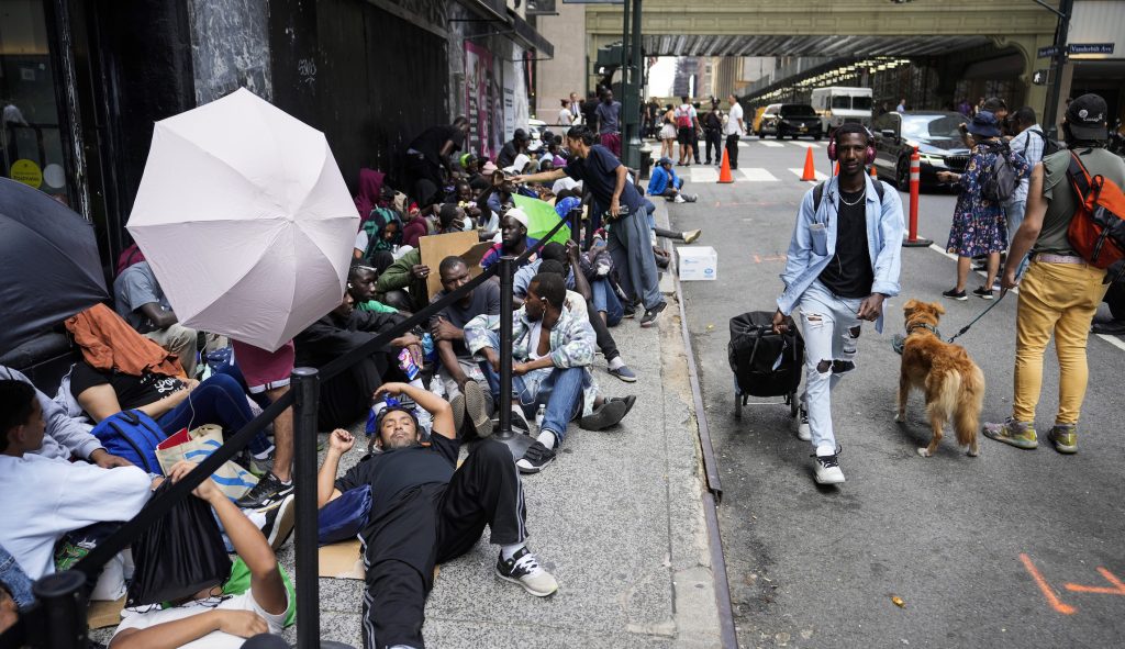 Immigrants sit in a queue outside of The Roosevelt Hotel that is being used by the city as temporary housing, Monday, July 31, 2023, in New York.